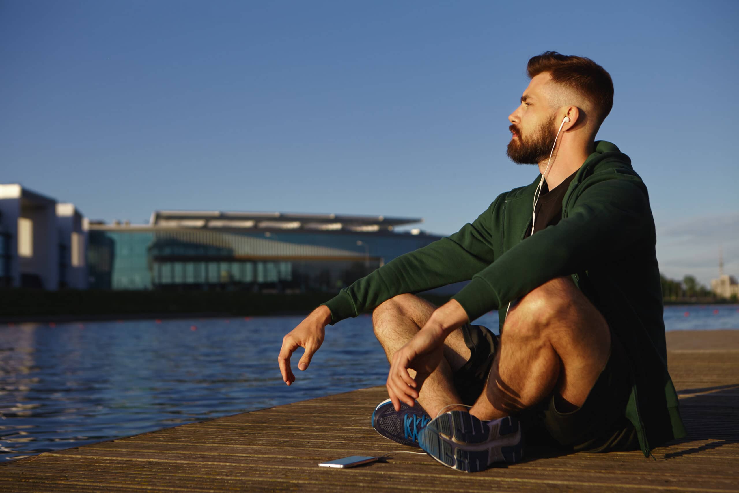 Bearded man in a green hoodie sits on a wooden dock by the water, wearing earbuds and looking to the right against a clear blue sky.